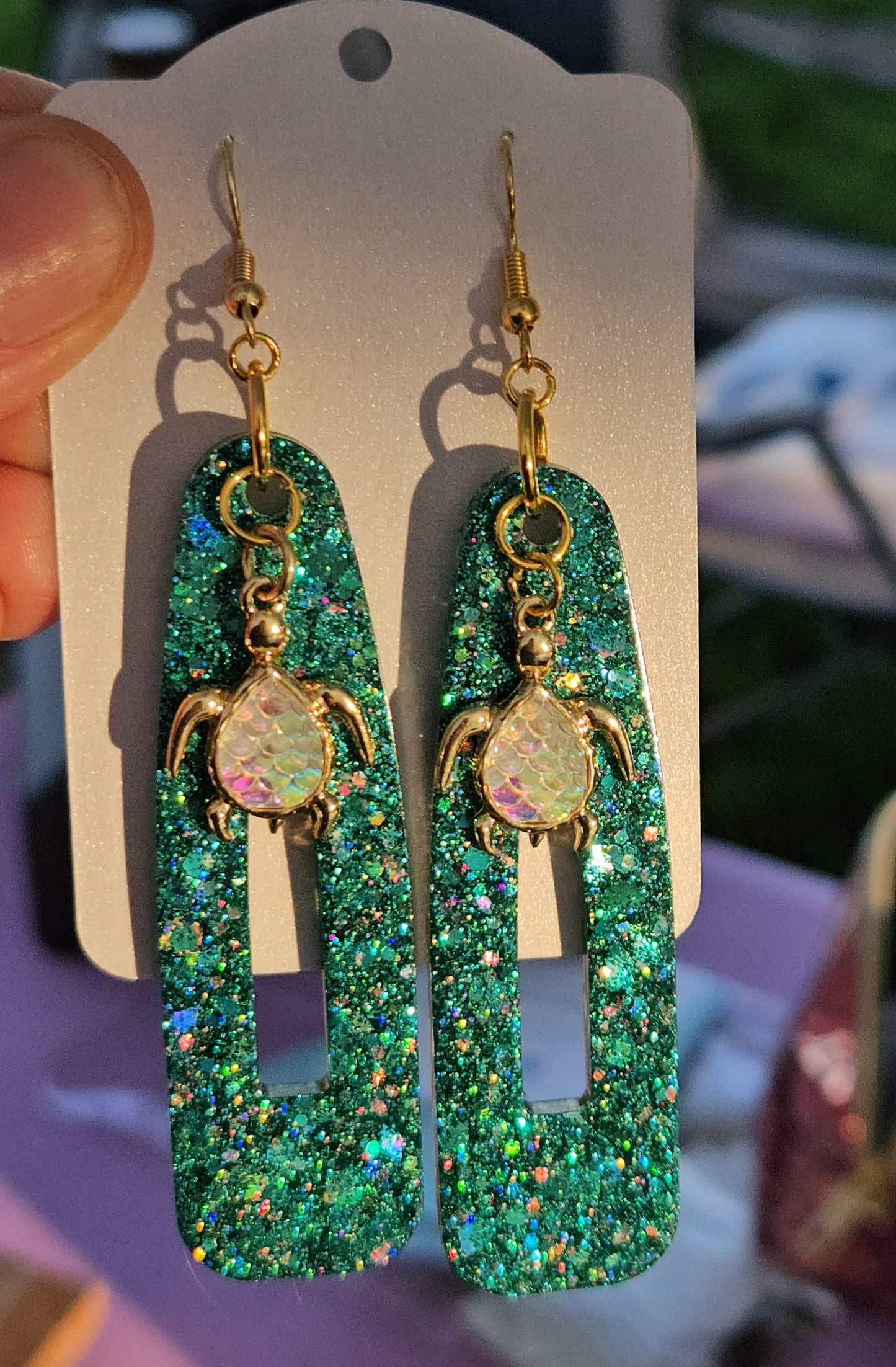 Green glittery earrings on a display card held by a hand with a blurred background